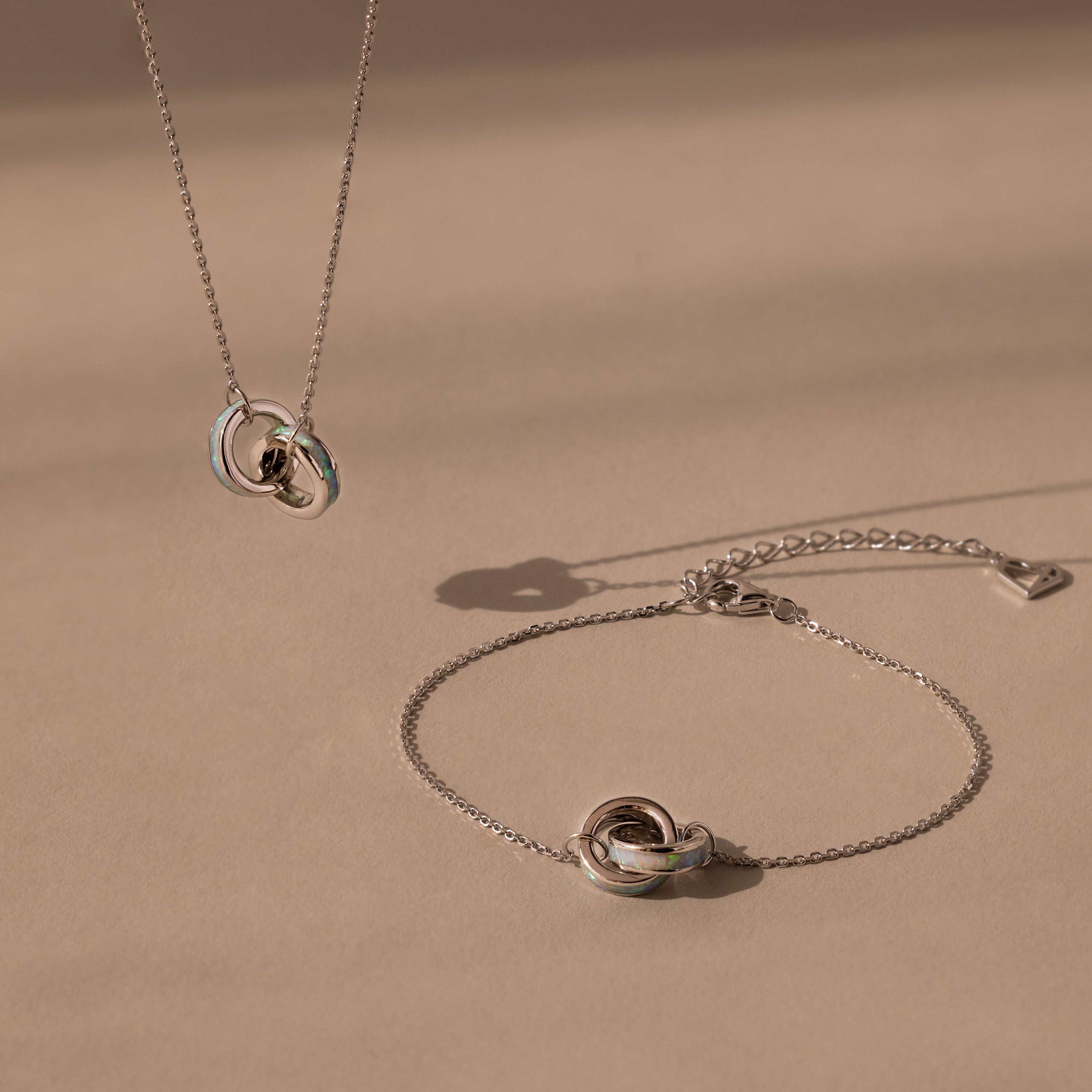 The Interlocking Opal Inlay Necklace and Bracelet Set in Silver features a knot pendant necklace with a matching bracelet, beautifully displayed against a neutral beige background.