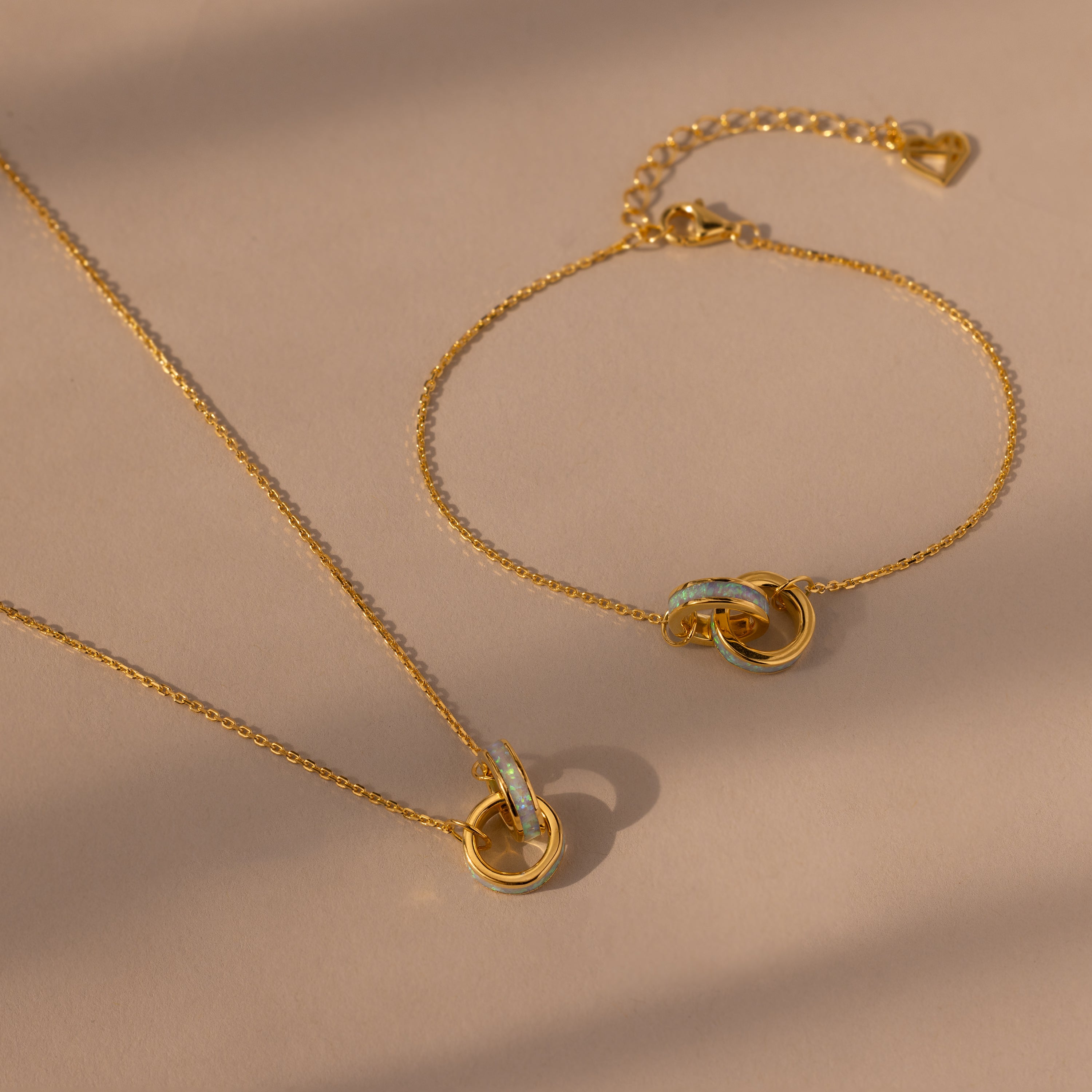 The Interlocking Opal Inlay Necklace and Bracelet Set in 18K Gold features delicate chains and interlocking rings, displayed on a beige background.