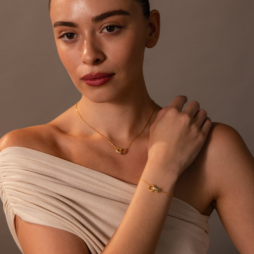A woman wears an Interlocking Opal Inlay Necklace and Bracelet Set in 18K Gold with an off-shoulder beige top, posing against a neutral background.