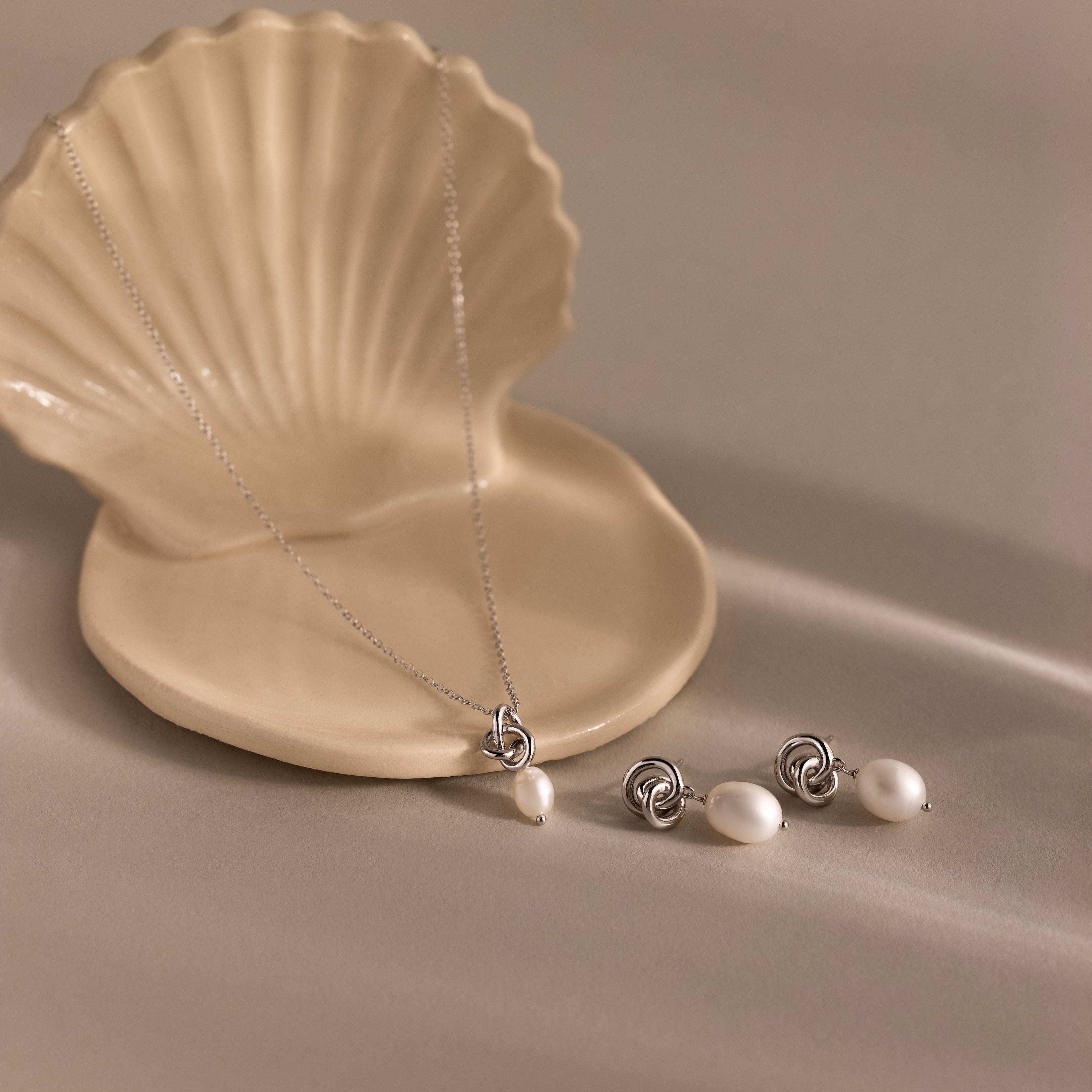 The Pearl Knot Necklace & Earrings Set in Sterling Silver is displayed on a beige shell-shaped dish against a neutral background.