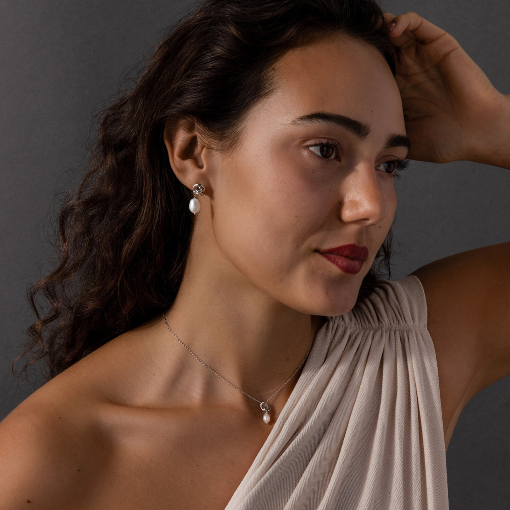 Woman with wavy dark hair wearing the Pearl Knot Necklace & Earrings Set and an off-white one-shoulder top—an elegant look, perfect as a gift for your wife.
