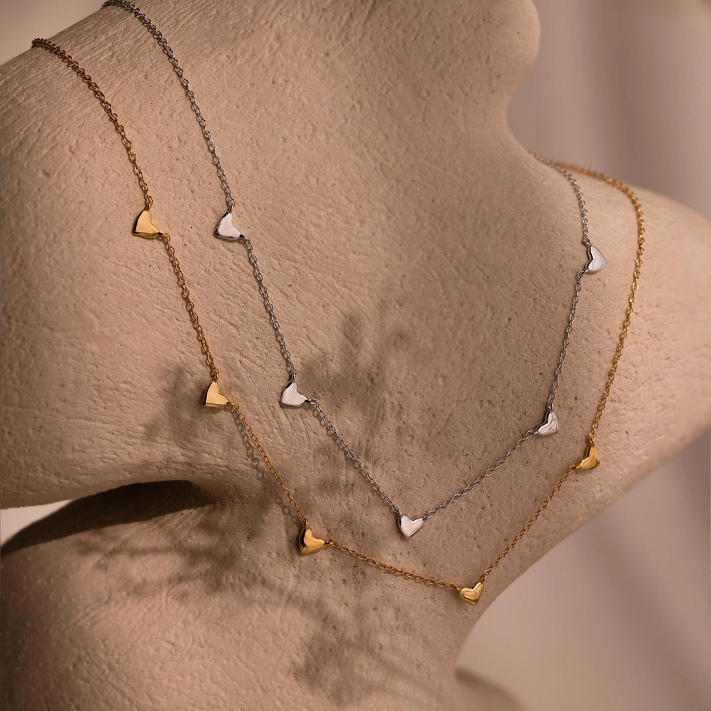 The Heart Station Necklace features delicate gold and silver heart charms, elegantly displayed on a beige textured bust.