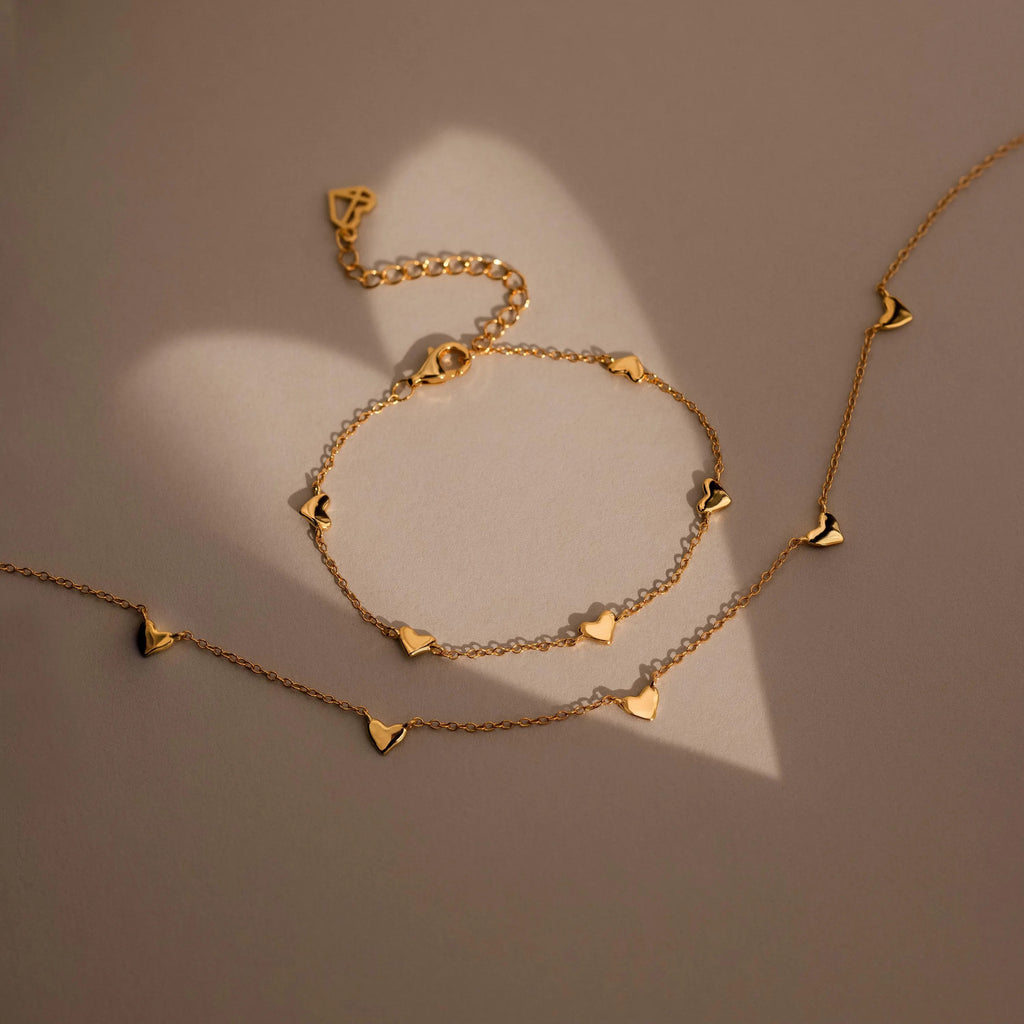 The Heart Station Necklace, a gold jewelry set with delicate heart charms, is displayed on a soft beige surface as natural light creates elegant heart-shaped shadows along its cable chain.