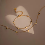 The Heart Station Necklace, a gold jewelry set with delicate heart charms, is displayed on a soft beige surface as natural light creates elegant heart-shaped shadows along its cable chain.
