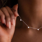 A person holds the Heart Station Necklace, featuring small heart charms and a fine cable chain, close to their neck.