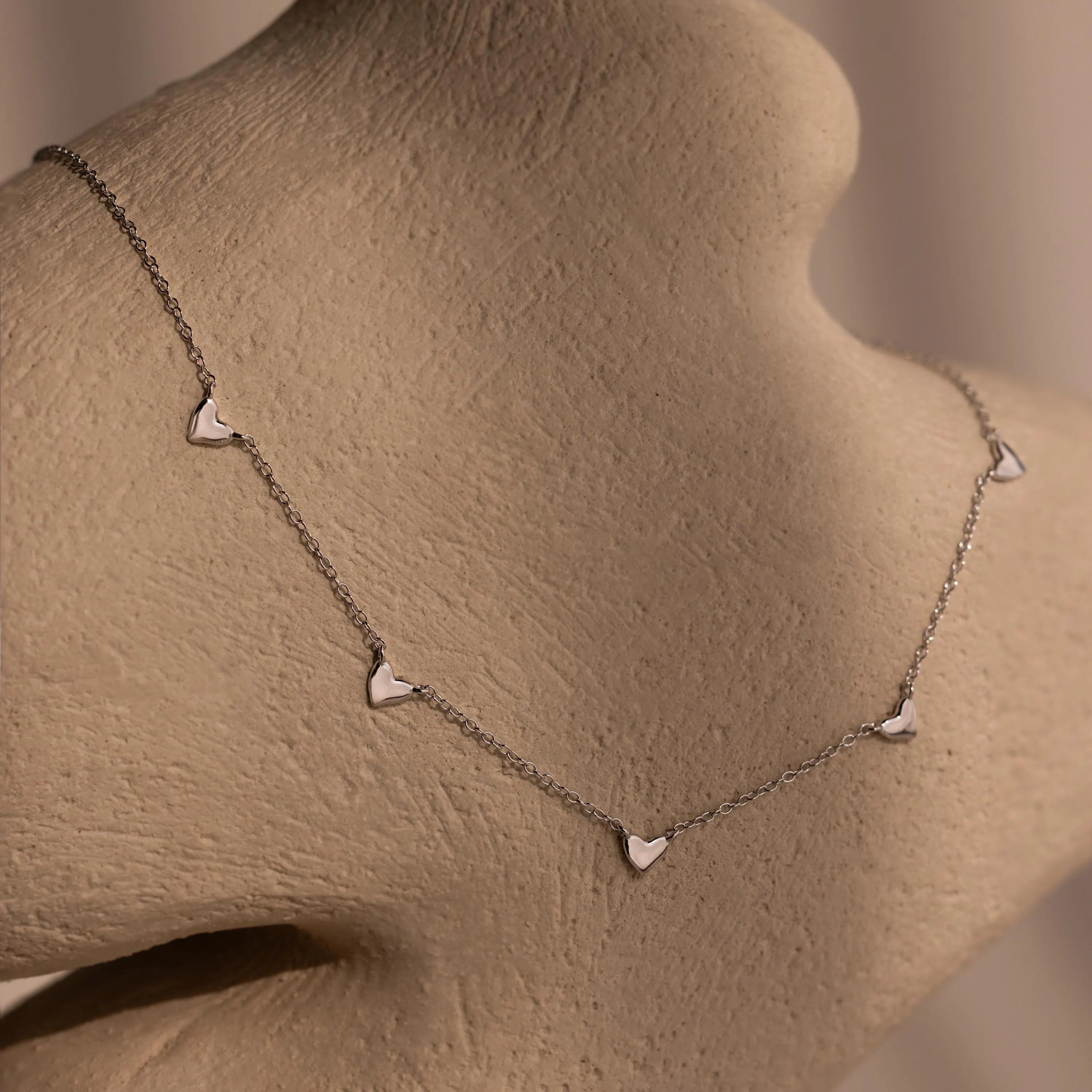 The Heart Station Necklace features small heart charms on a delicate cable chain, shown on a beige textured mannequin bust.