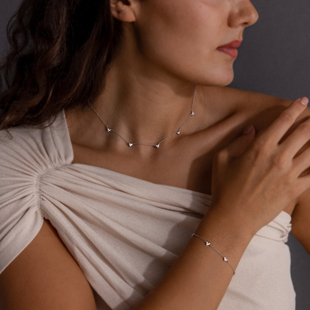 A woman wearing a cream off-shoulder top and the Heart Station Necklace & Bracelet Set against a dark background.