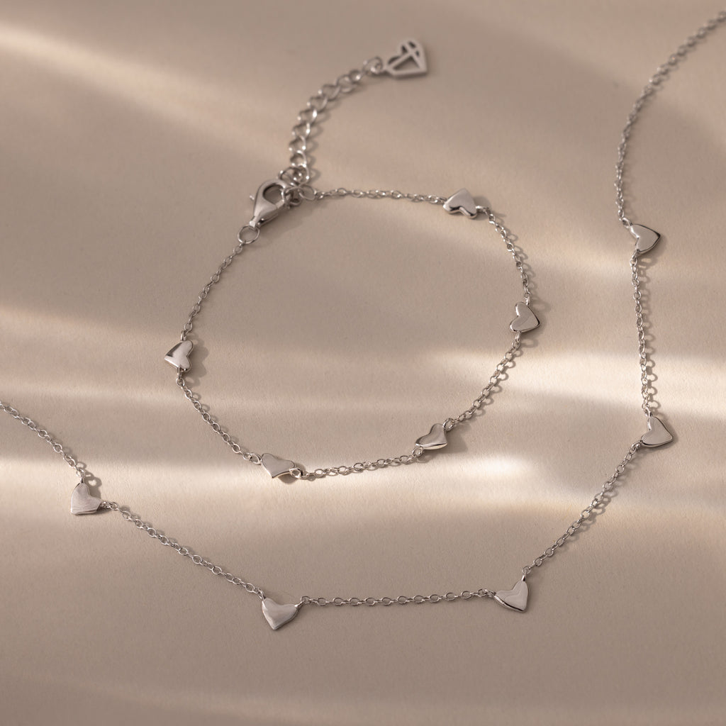 Heart Station Necklace & Bracelet Set in Sterling Silver featuring small heart-shaped charms displayed on a beige surface with soft lighting.