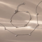 Heart Station Necklace & Bracelet Set in Sterling Silver featuring small heart-shaped charms displayed on a beige surface with soft lighting.