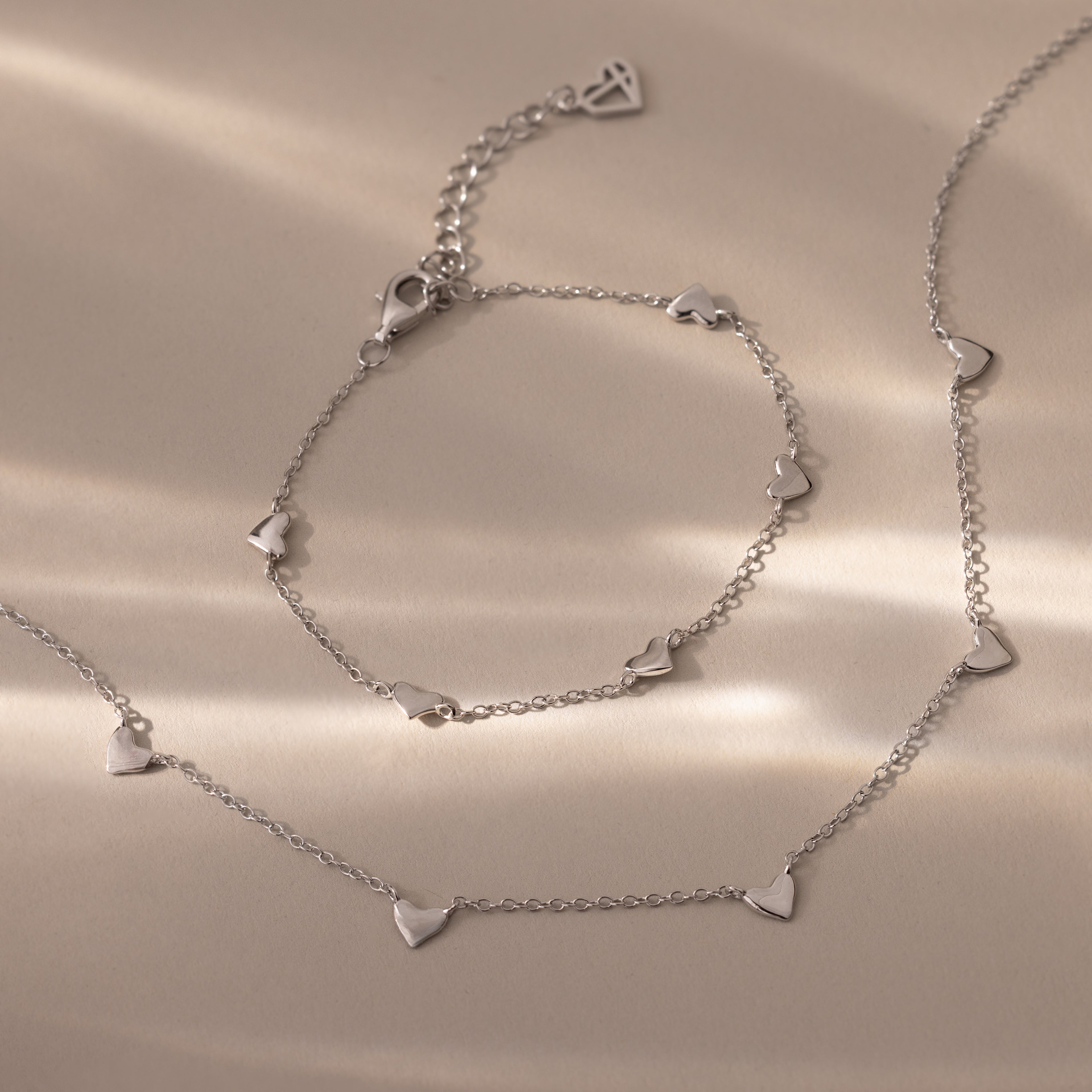 Heart Station Necklace & Bracelet Set in Sterling Silver featuring small heart-shaped charms displayed on a beige surface with soft lighting.