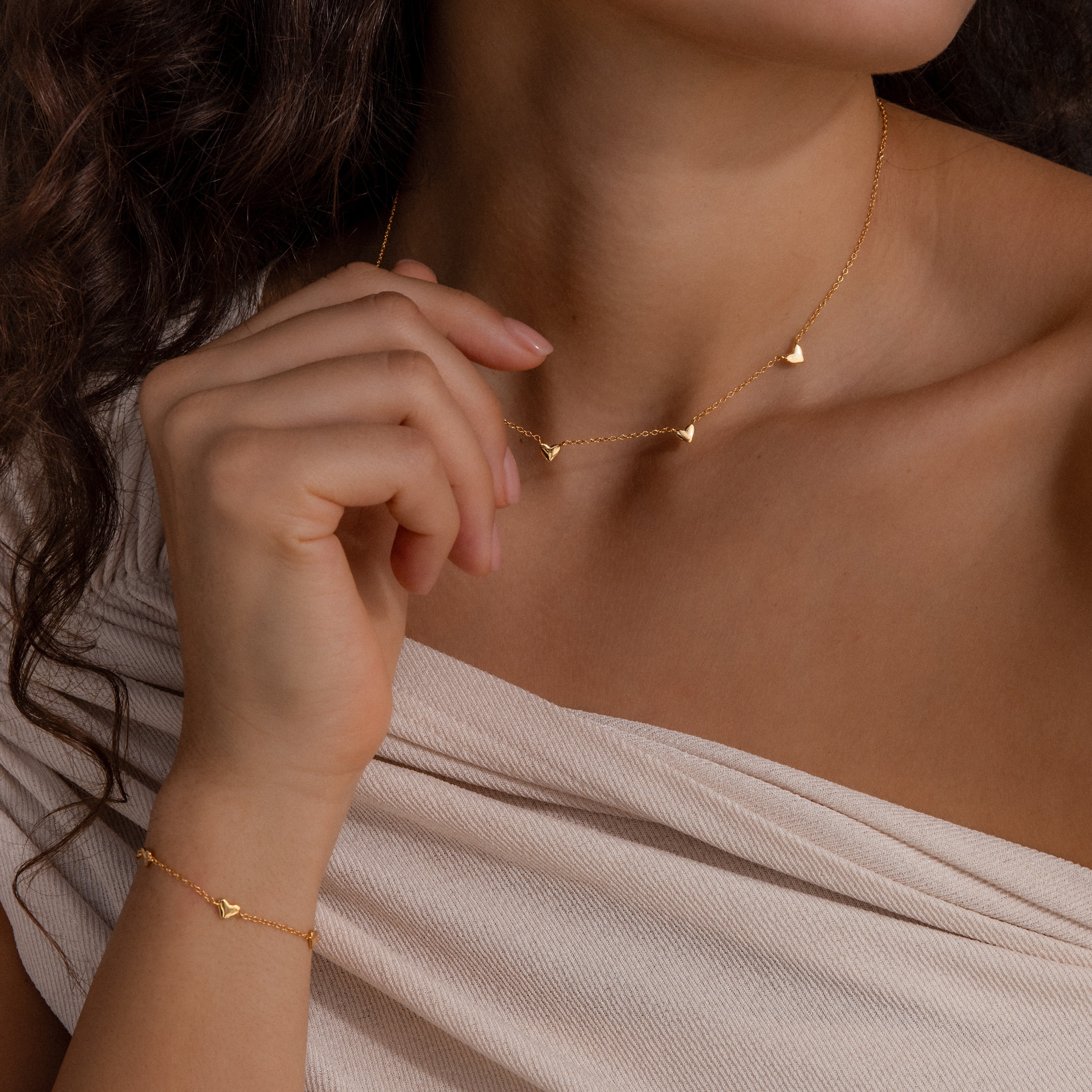 A woman in a light off-shoulder top wears the Heart Station Necklace & Bracelet Set, featuring small heart charms.