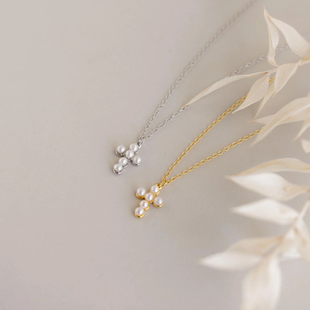 Two Pearl Cross Necklaces display pearl cross charms on gold and silver chains, shown on a light surface with dried leaves.