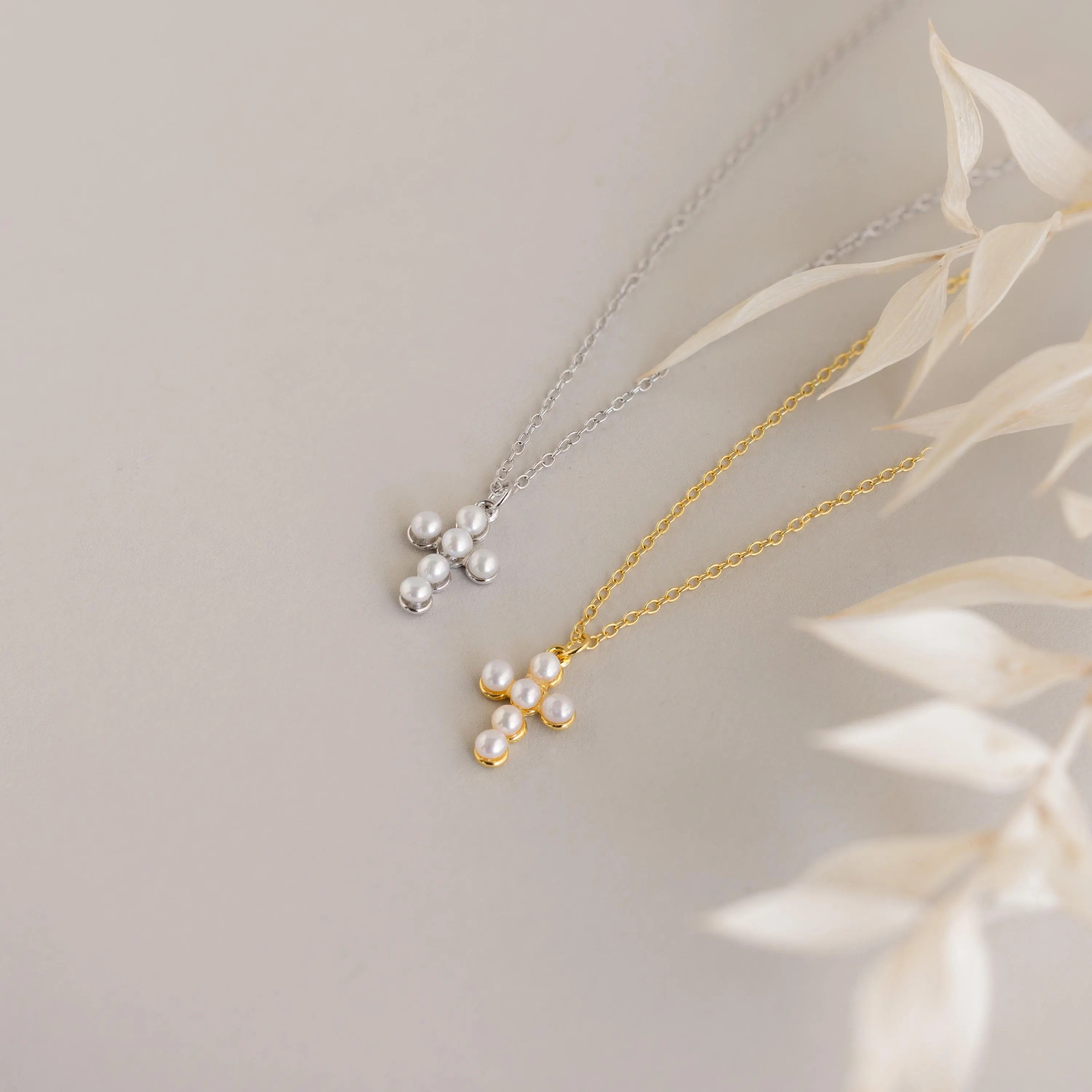 Two Pearl Cross Necklaces display pearl cross charms on gold and silver chains, shown on a light surface with dried leaves.