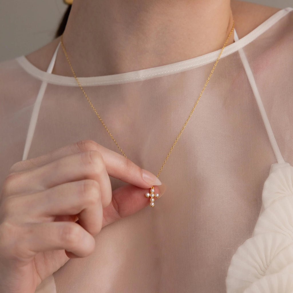 A woman in a sheer white top holds the Pearl Cross Necklace, featuring a delicate gold chain and a cross charm adorned with luminous pearls.