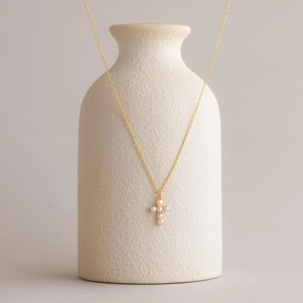 The Pearl Cross Necklace, featuring a radiant pearl cross pendant, is showcased on a textured white ceramic vase.