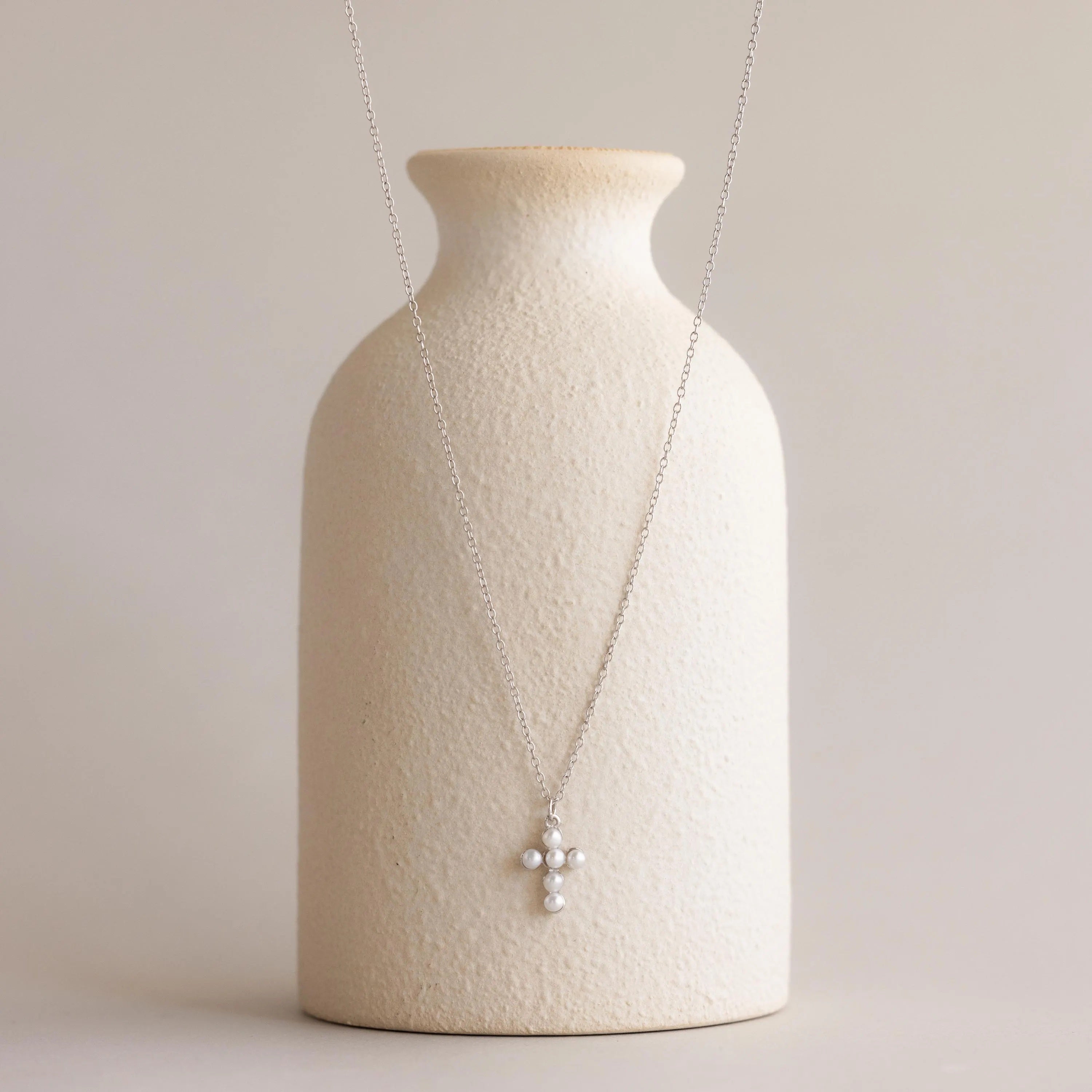 Pearl Cross Necklace adorned with luminous pearls, elegantly displayed on a textured cream vase.