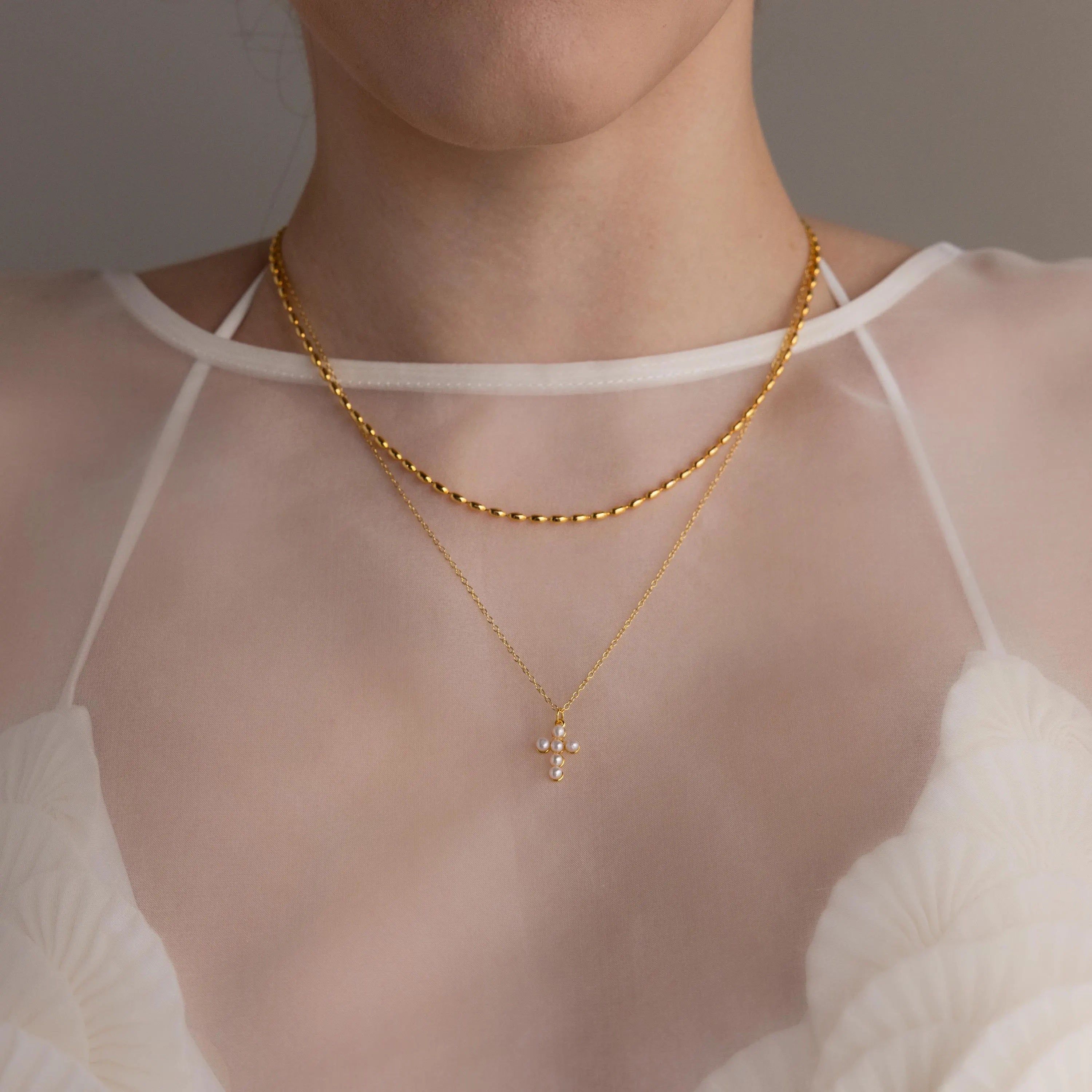 Close-up of a woman wearing two gold necklaces, one of which is the Pearl Cross Necklace, styled over a sheer white floral top.