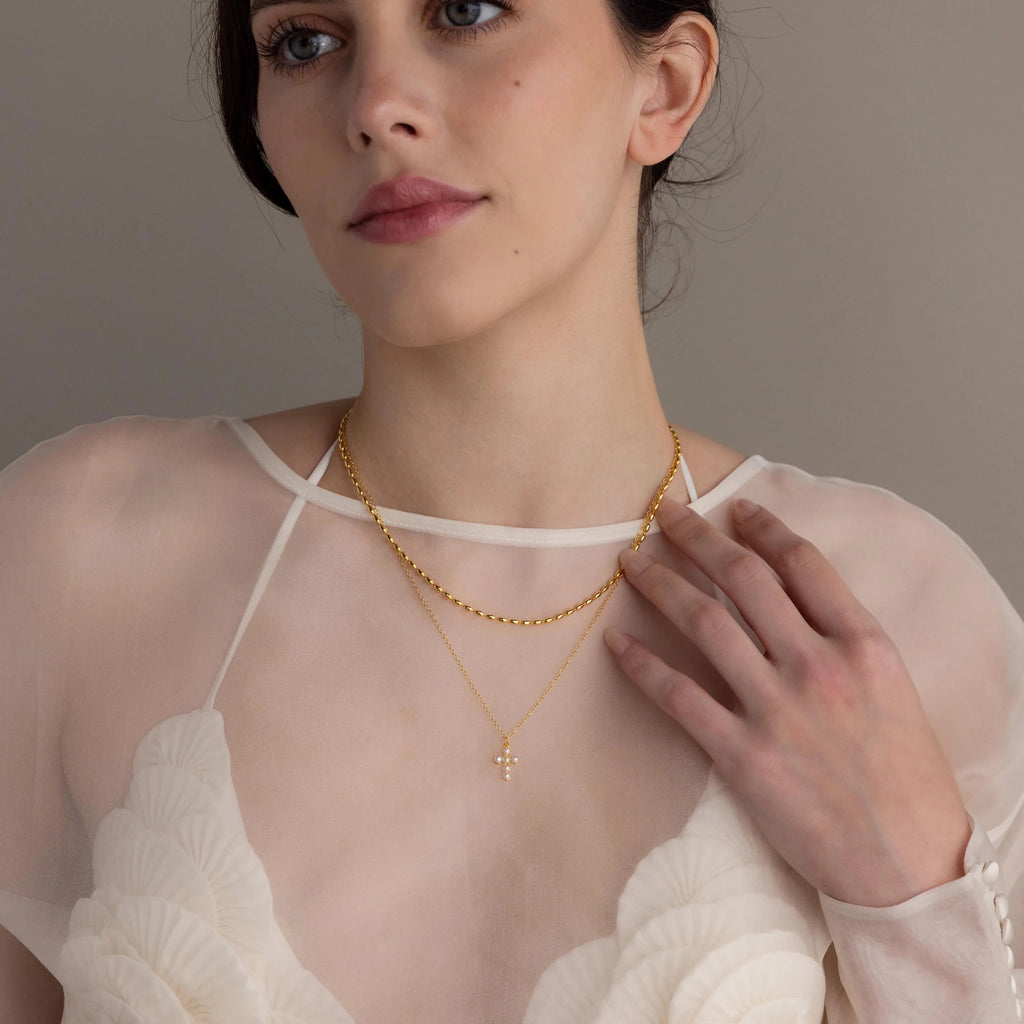 A woman in a sheer white top wears the Pearl Cross Necklace—two gold chains, one with a delicate cross charm and the other with luminous pearls—as she gazes thoughtfully to the side.