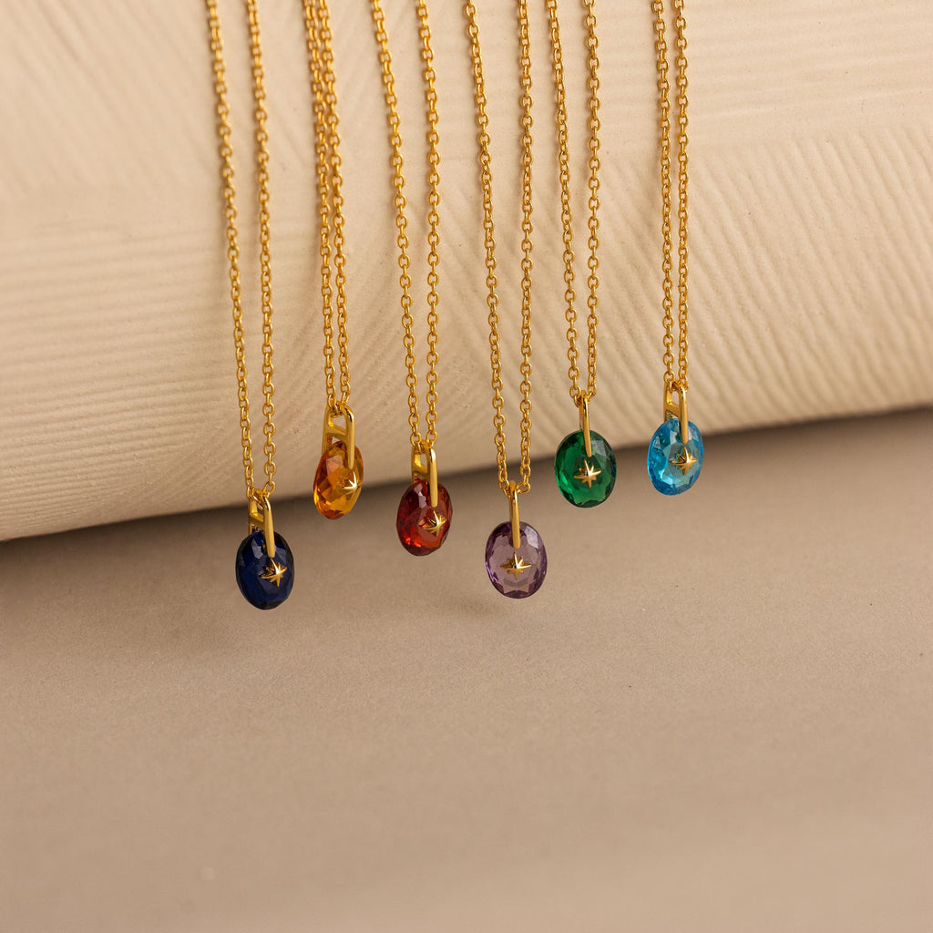 6 Gold North Star Birthstone Pendant Necklaces displayed hanging over a soft beige surface.
