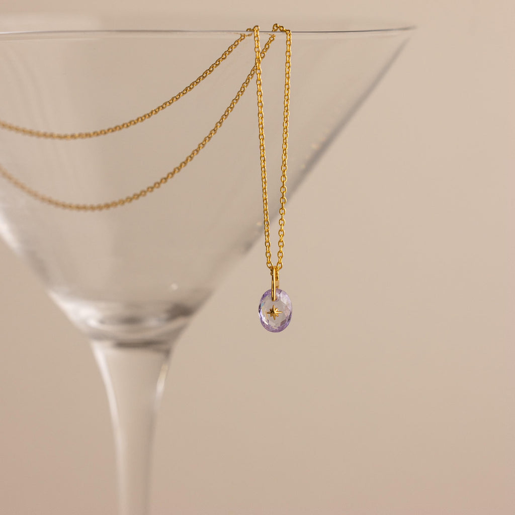 Gold North Star Birthstone Necklace with a Light Purple Alexandrite Gemstone hanging inside a cocktail glass.