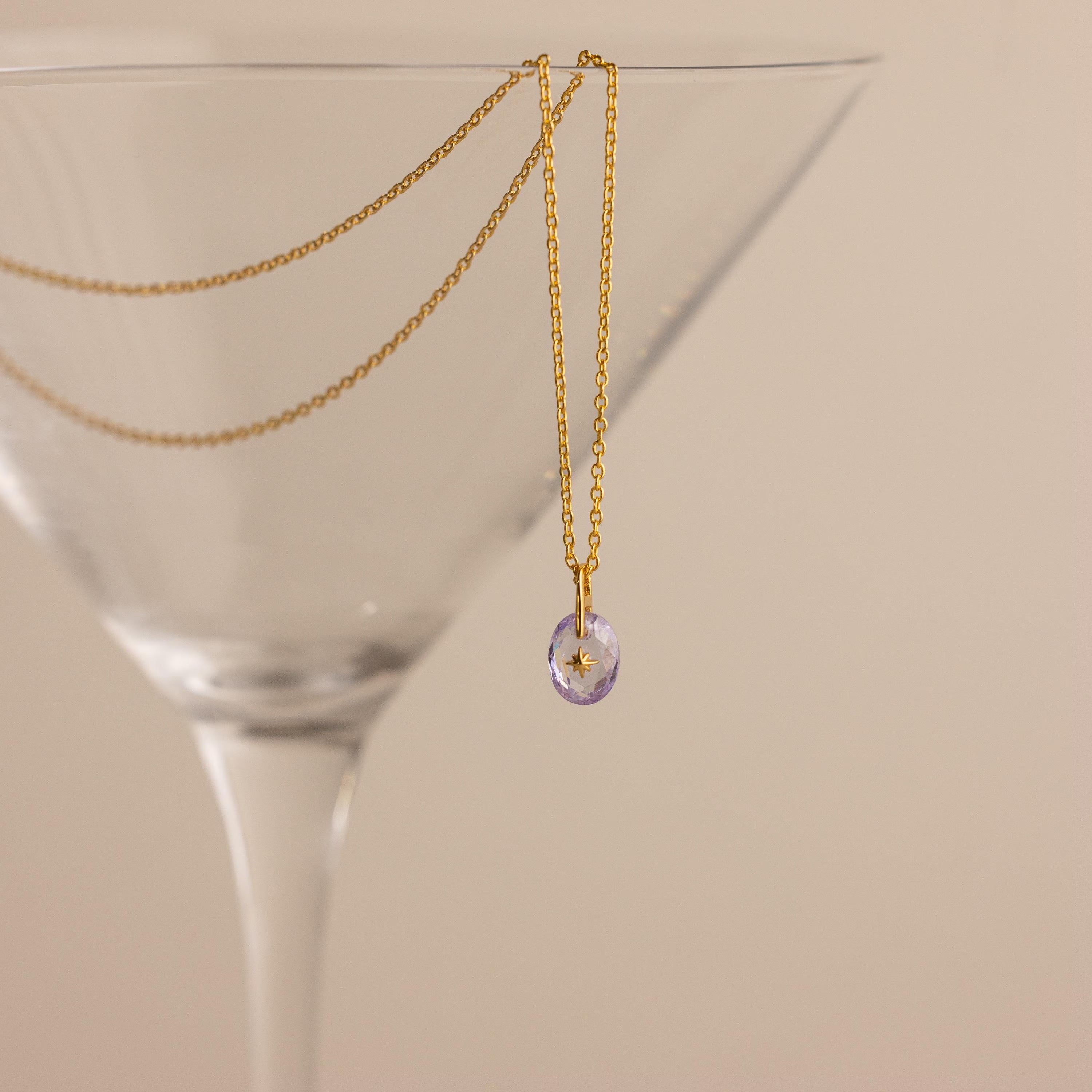 Gold North Star Birthstone Necklace with a Light Purple Alexandrite Gemstone hanging inside a cocktail glass.