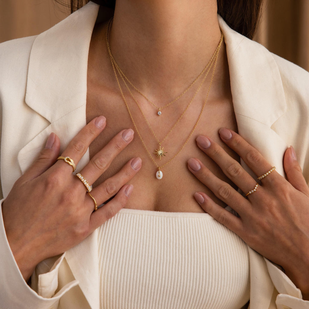 Woman wearing the North Star Birthstone Signet Necklace in Garnet, layered with other gold jewelry, a cream blazer, and a ribbed white top, with her hands near her chest.