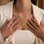 Woman wearing the North Star Birthstone Signet Necklace in Garnet, layered with other gold jewelry, a cream blazer, and a ribbed white top, with her hands near her chest.