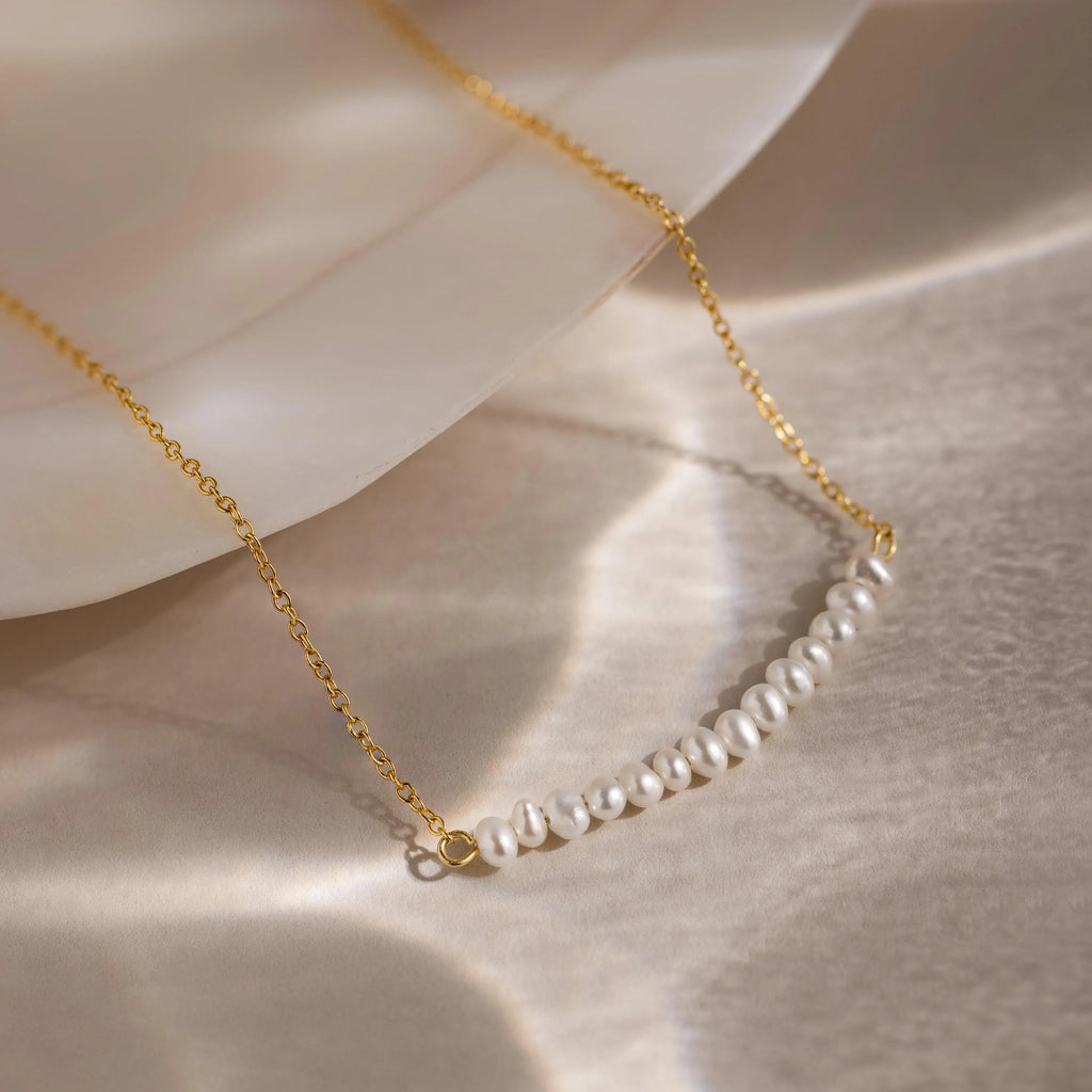 The Pearl Curve Necklace, featuring a gold chain with small white pearls, is perfect as bridal jewelry and is displayed on a light, reflective surface beside a shell.
