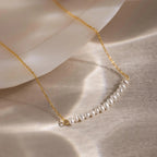 The Pearl Curve Necklace, featuring a gold chain with small white pearls, is perfect as bridal jewelry and is displayed on a light, reflective surface beside a shell.