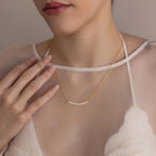 A woman wearing a sheer white top gently touches her collarbone, showcasing the delicate Pearl Curve Necklace—perfect as bridal jewelry.