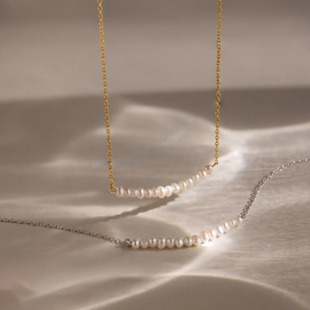Two Pearl Curve Necklaces, ideal for bridal jewelry, are displayed on a beige surface—one features a gold chain and the other a silver chain.