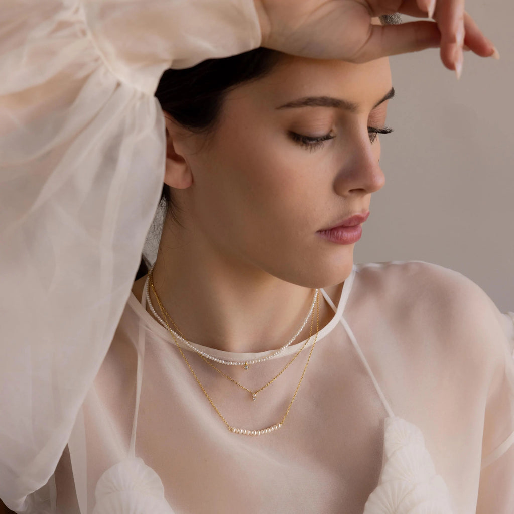 Woman in sheer white top wearing layered gold necklaces and the Pearl Curve Necklace—a delicate accent for a graceful, modern bridal jewelry look.