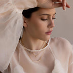 Woman in sheer white top wearing layered gold necklaces and the Pearl Curve Necklace—a delicate accent for a graceful, modern bridal jewelry look.