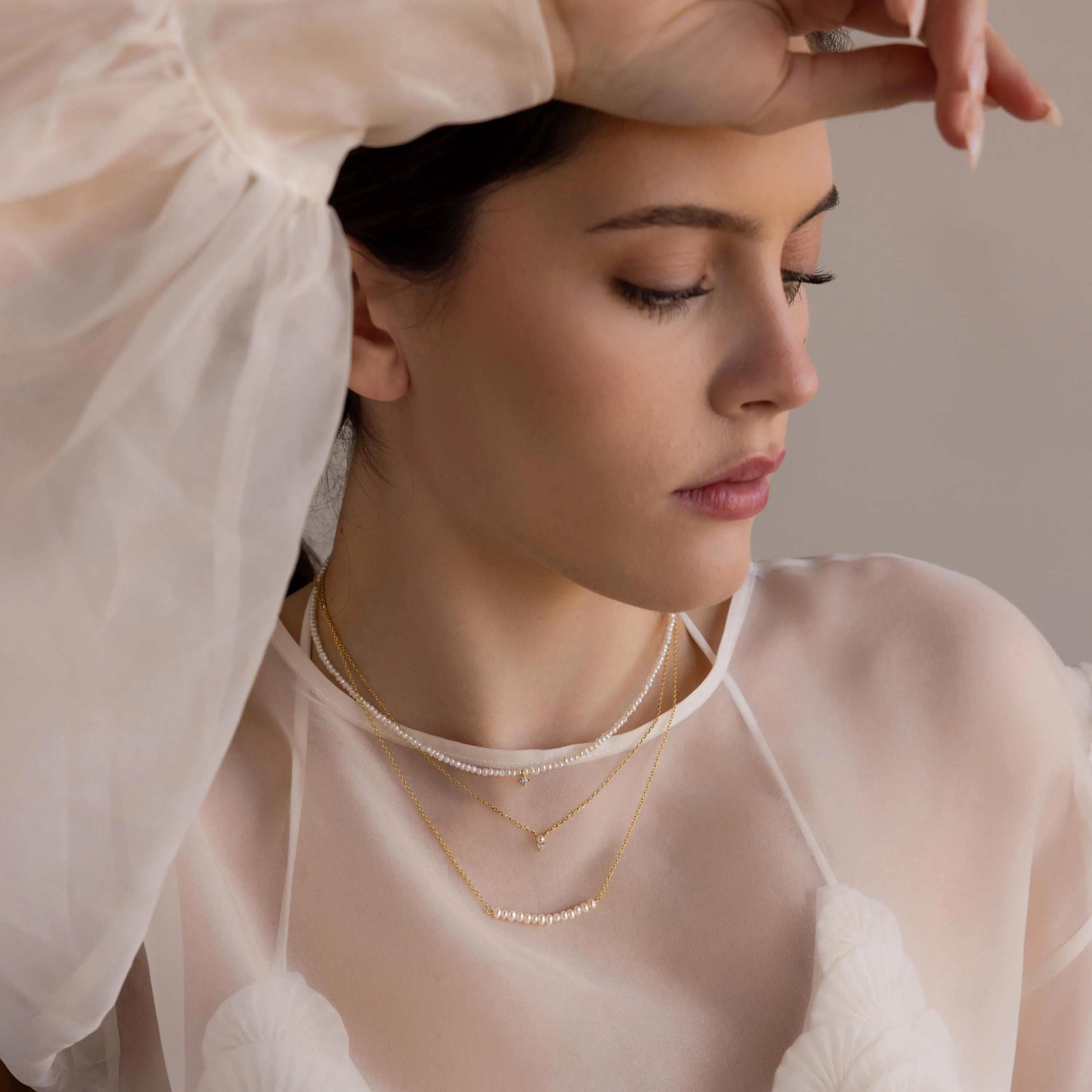 Woman in sheer white top wearing layered gold necklaces and the Pearl Curve Necklace—a delicate accent for a graceful, modern bridal jewelry look.