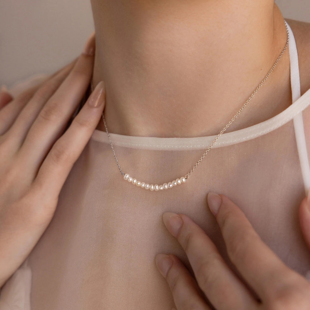A woman wears the Pearl Curve Necklace with a sheer blouse, touching her collarbone with manicured hands—a perfect choice for elegant wedding jewelry.