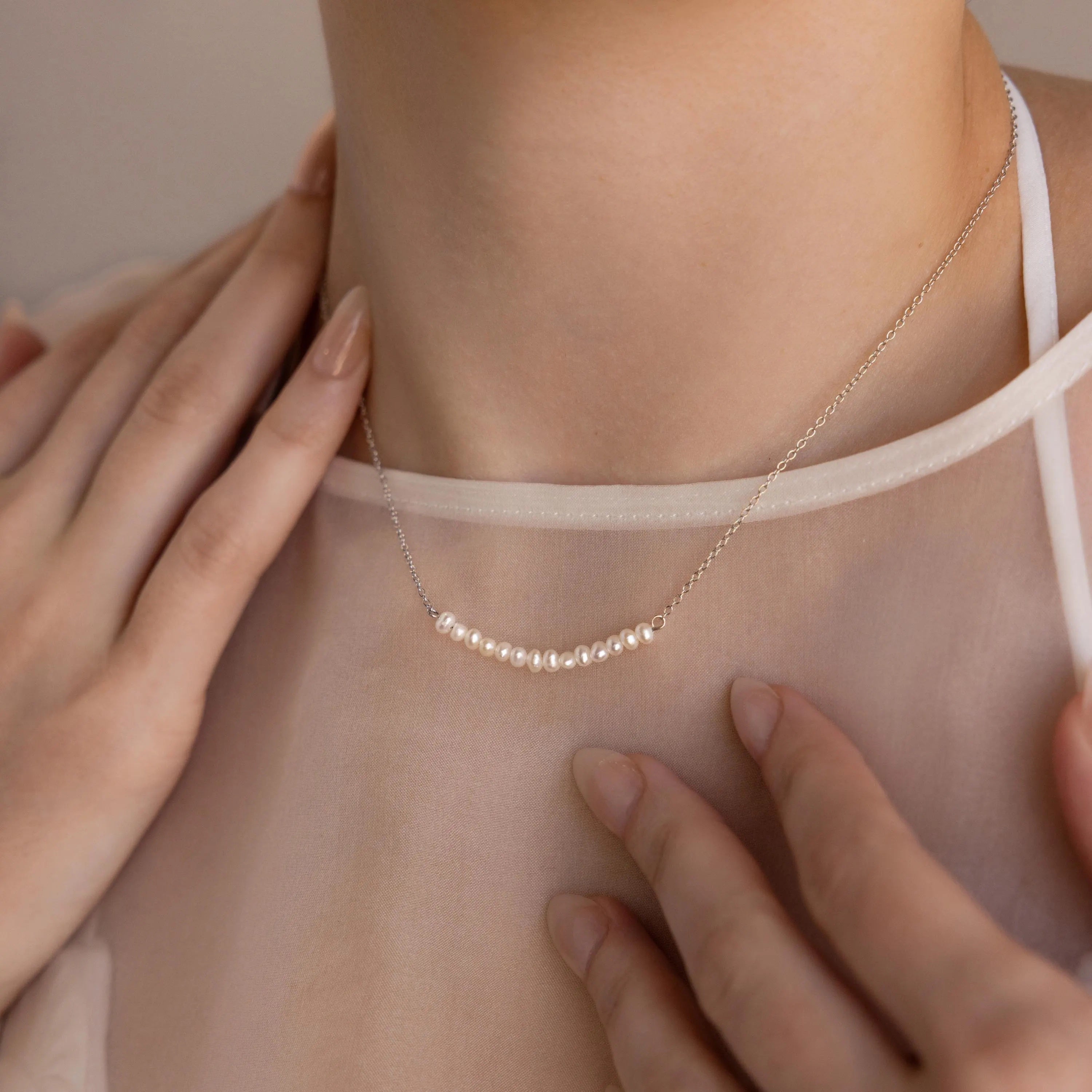 A woman wears the Pearl Curve Necklace with a sheer blouse, touching her collarbone with manicured hands—a perfect choice for elegant wedding jewelry.
