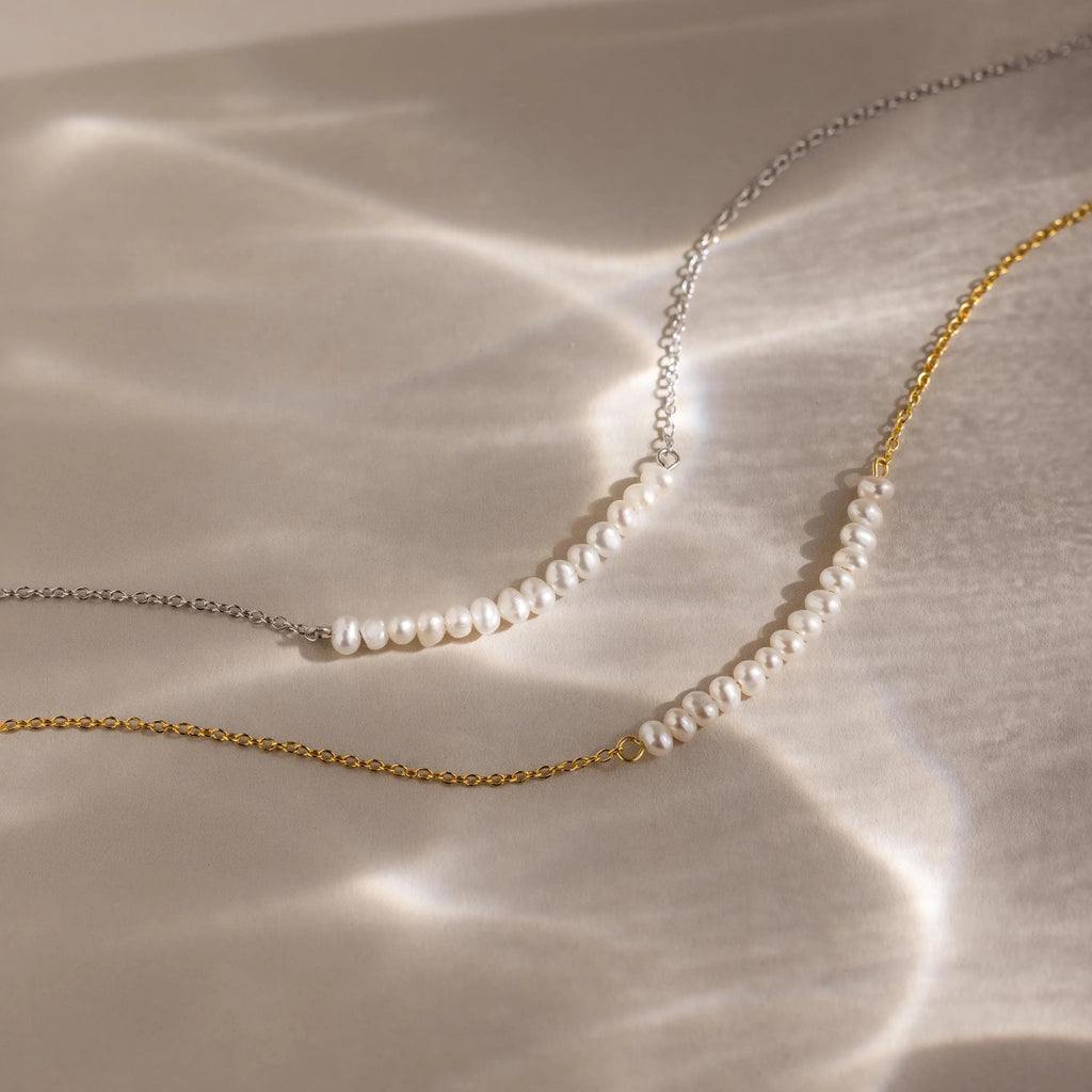 The Pearl Curve Necklace, available on gold and silver chains, is elegantly displayed on a reflective surface—an ideal choice for bridal jewelry.