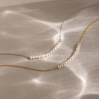The Pearl Curve Necklace, available on gold and silver chains, is elegantly displayed on a reflective surface—an ideal choice for bridal jewelry.