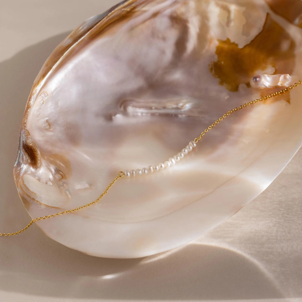 The Pearl Curve Necklace, featuring a delicate gold chain and lustrous pearls, rests inside a shiny, iridescent seashell on a beige surface—an elegant choice for bridal jewelry.