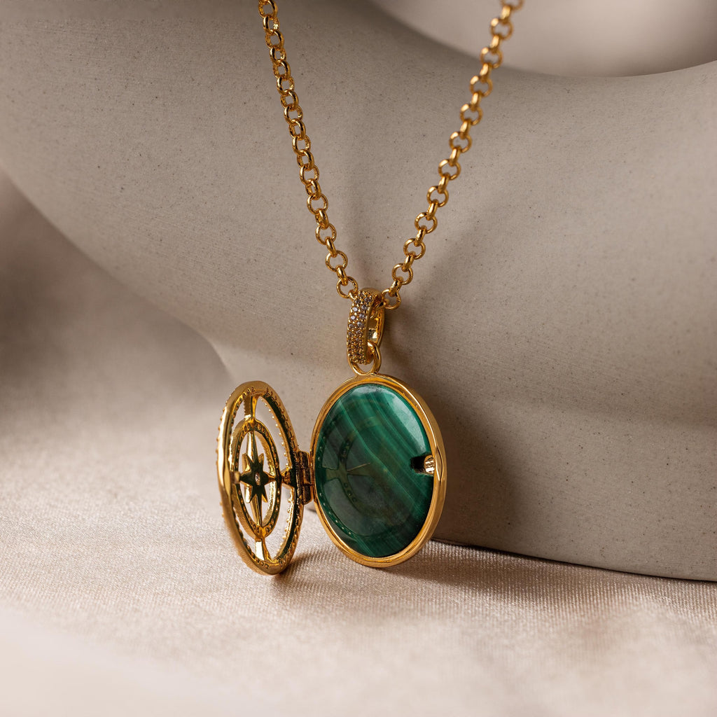 The Malachite Compass Locket Necklace, featuring a green malachite stone and a partially open locket, rests on smooth beige fabric—a distinctive guidance necklace inspired by adventure.
