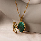 The Malachite Compass Locket Necklace, featuring a green malachite stone and a partially open locket, rests on smooth beige fabric—a distinctive guidance necklace inspired by adventure.