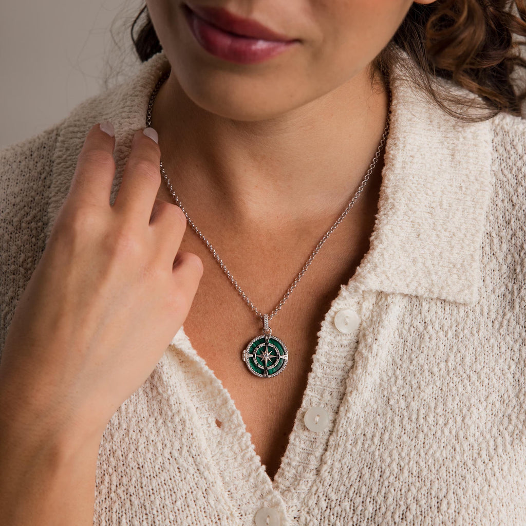 A woman touches her collar while wearing a cream textured button-up top and the Malachite Compass Locket Necklace.