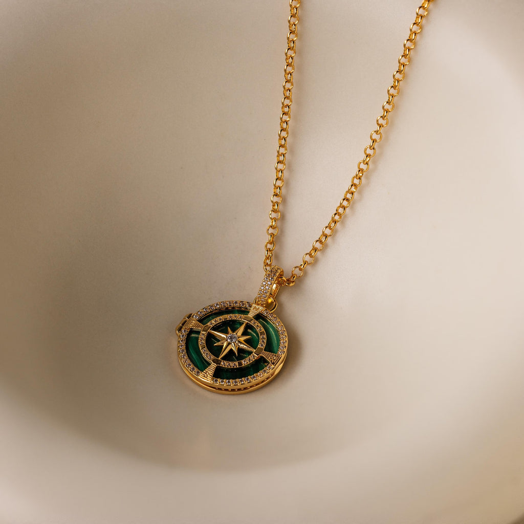 The Malachite Compass Locket Necklace features a gold chain and a green-and-gold compass pendant, displayed on a cream surface—a stylish guidance necklace perfect for any occasion.