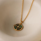 The Malachite Compass Locket Necklace features a gold chain and a green-and-gold compass pendant, displayed on a cream surface—a stylish guidance necklace perfect for any occasion.