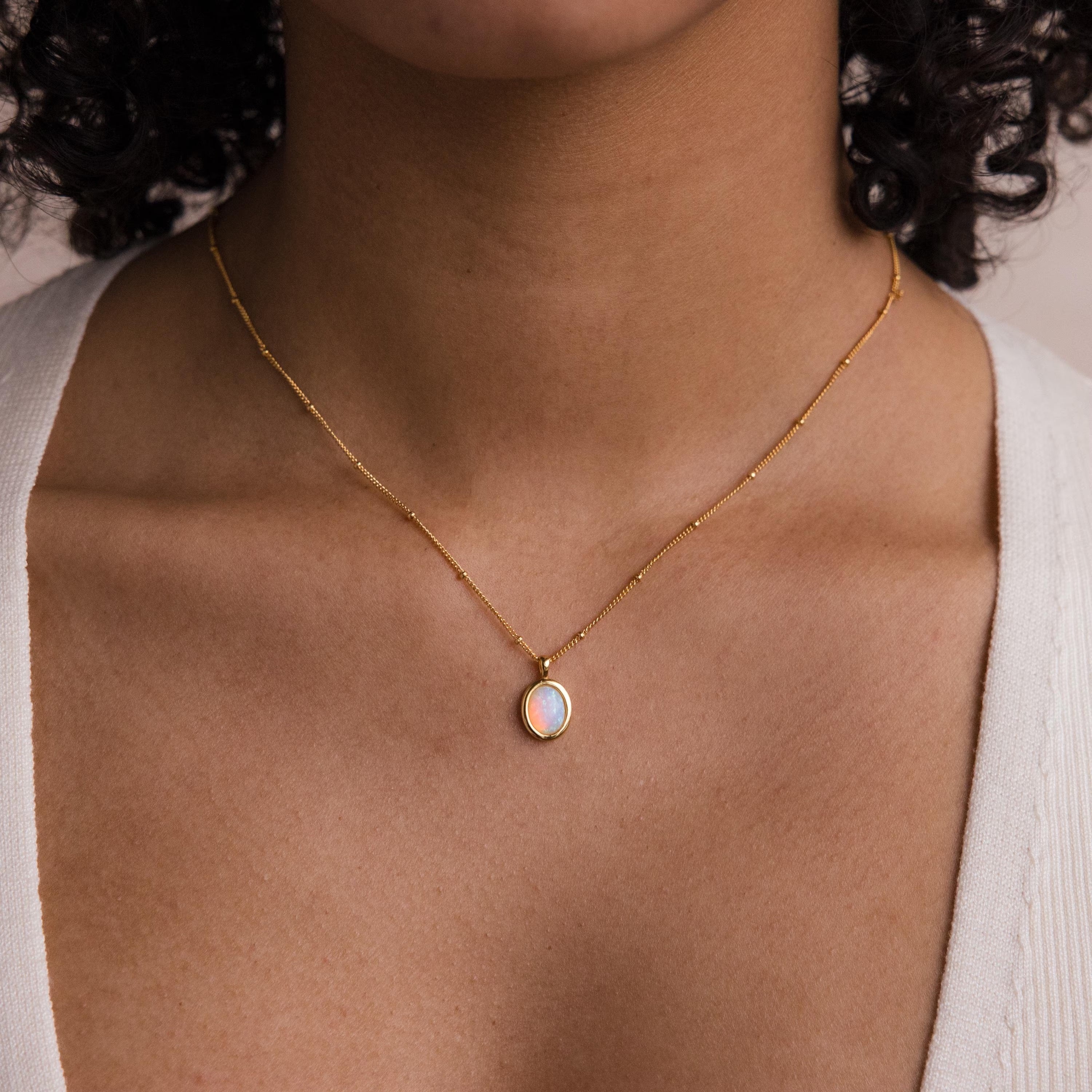 A woman shown from the shoulders up wears the Opal Pendant Necklace, featuring a gold satellite chain with an oval opal gemstone.