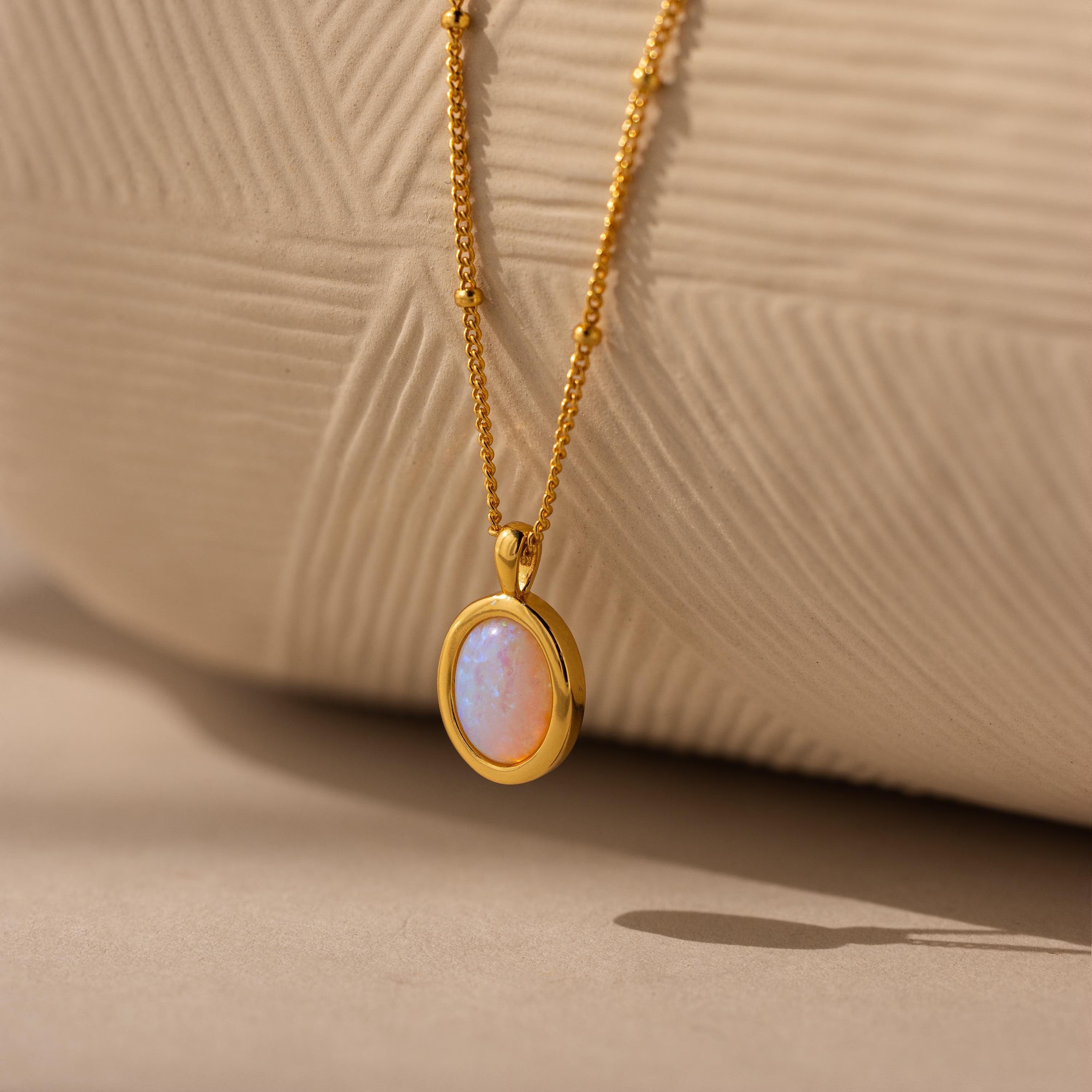 Opal Pendant Necklace: Showcasing an oval Opal gemstone on a fine gold satellite chain, beautifully presented against a textured beige background.