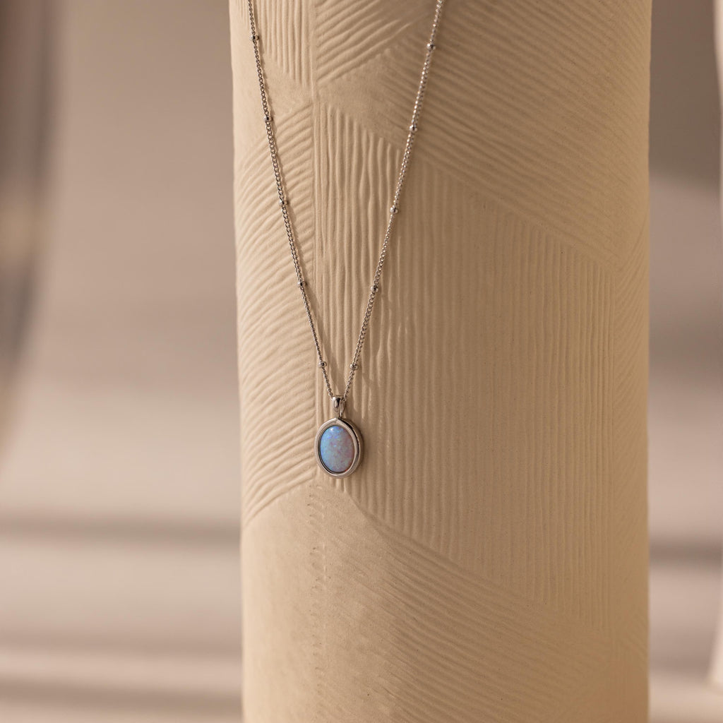 Opal Pendant Necklace with a small round opal gemstone, shown on a textured beige cylindrical surface.