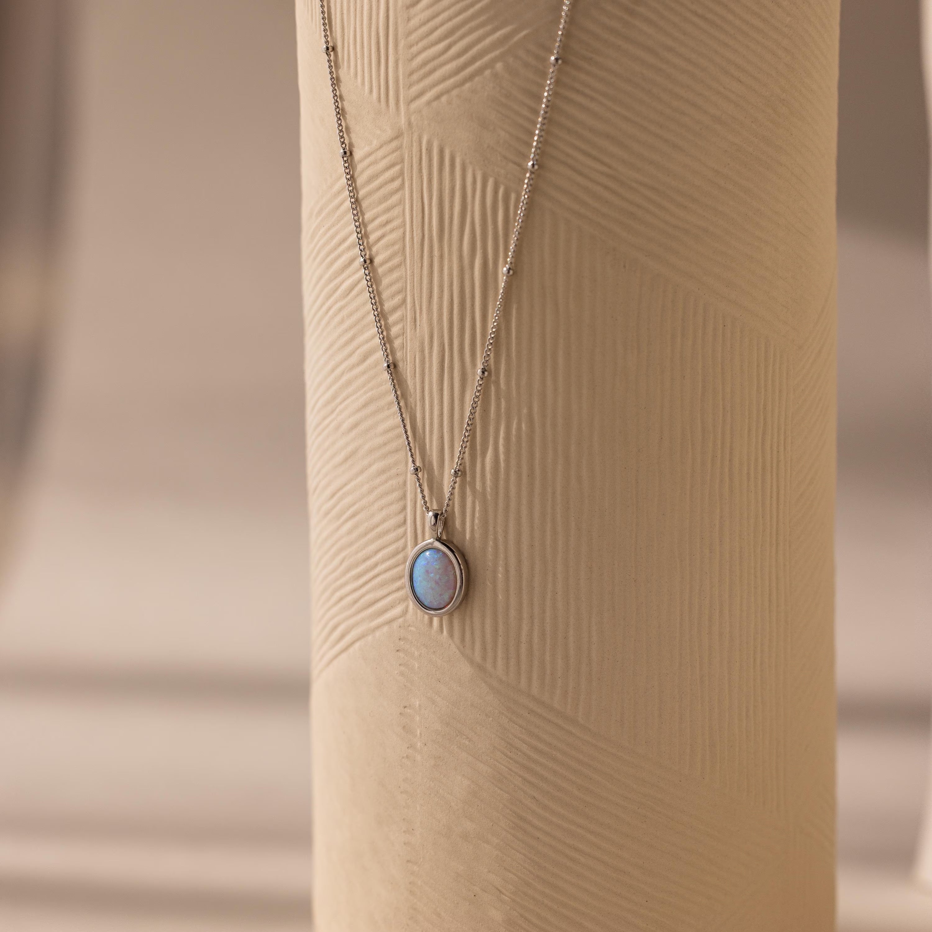 Opal Pendant Necklace with a small round opal gemstone, shown on a textured beige cylindrical surface.