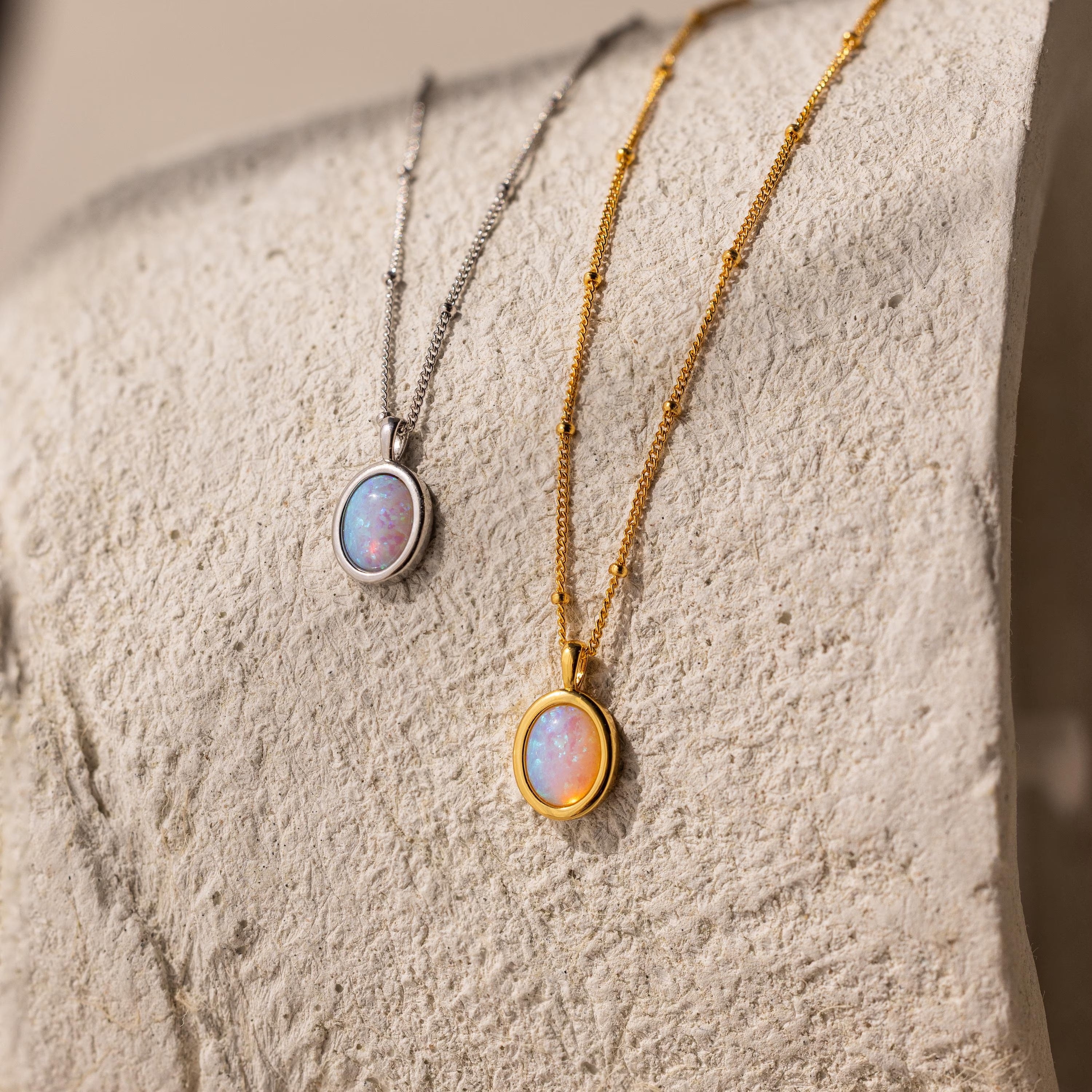 Two Opal Pendant Necklaces are shown on a textured beige mannequin stand—both featuring dazzling Opal gemstones, one with a silver satellite chain and the other with a gold chain.