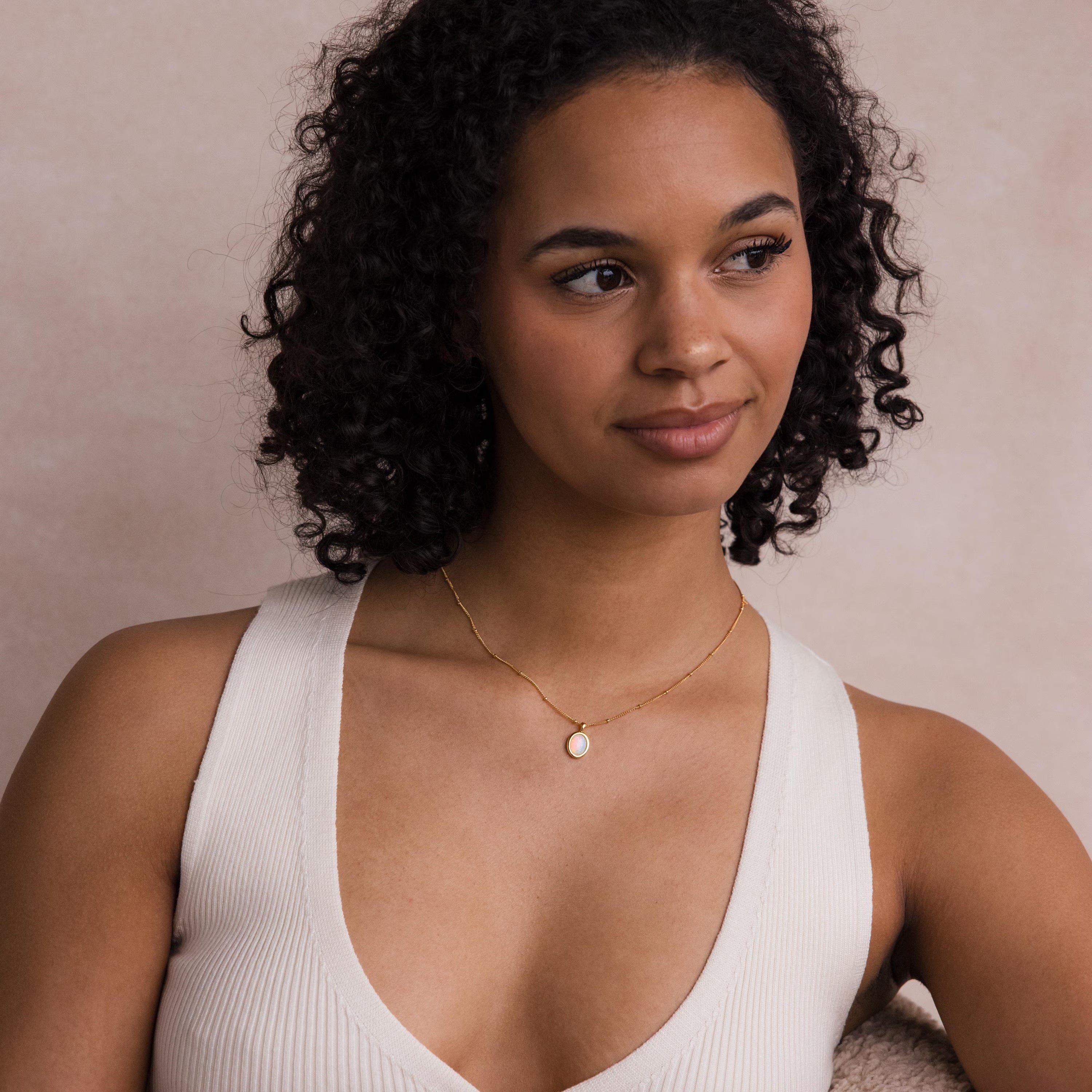 A woman with curly hair, wearing a white top and the Opal Pendant Necklace on a delicate satellite chain, gazes softly to the side.